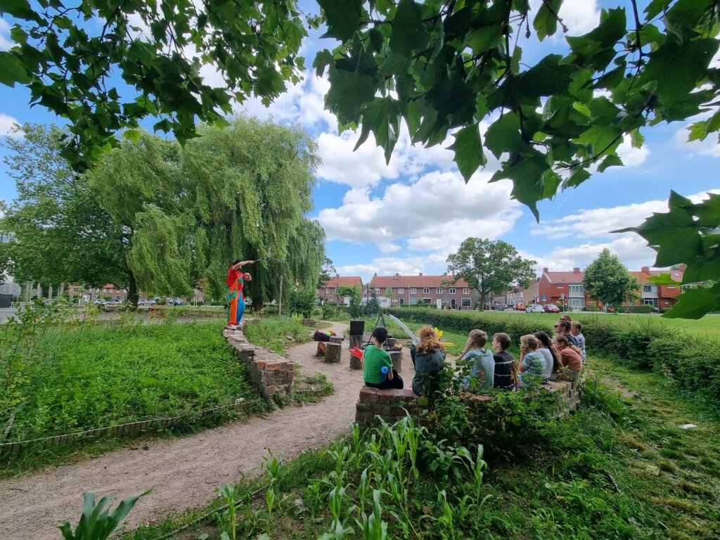 Een middenberm is omgevormd tot plek om samen te komen. Er is een ronde muur aangelegd. Op de foto zie je kinderen zitten op de muur. Ze kijken naar een optreden van een vrolijk geklede man die een ballontruc laat zien. 